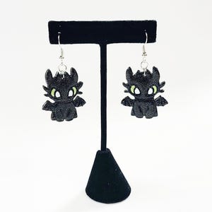 May include: A pair of black dangle earrings featuring a cartoon dragon with green eyes. The dragon is made of a sparkly material and is hanging from silver hooks.