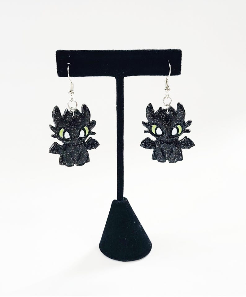 May include: A pair of black dangle earrings featuring a cartoon dragon with green eyes. The dragon is made of a sparkly material and is hanging from silver hooks.