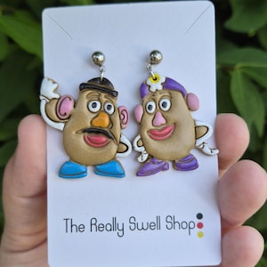 May include: A pair of dangle earrings featuring two potato-shaped characters with plastic stud posts. One character wears a black hat, blue shoes, and a mustache. The other wears a purple hat with a flower, pink ears, and purple shoes. Text reads The Really Swell Shop.
