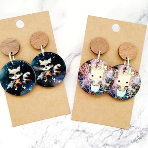 May include: Two pairs of dangle earrings with wooden tops. One pair features a cartoon raccoon with a gun, the other pair features a cartoon tree creature. Both pairs have a galaxy background.