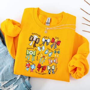 May include: Mustard yellow sweatshirt featuring colorful bow designs representing various animated movies, including "Ratatouille," "Wall-E," and "Cars." The sweatshirt has a crew neck and long sleeves. A gold necklace with a pendant is visible.