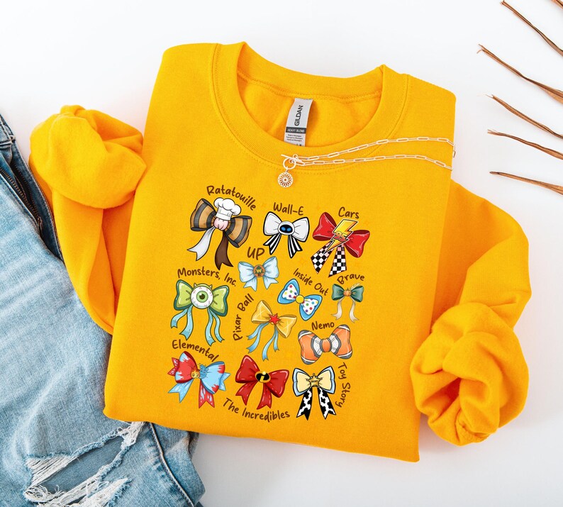 May include: Mustard yellow sweatshirt featuring colorful bow designs representing various animated movies, including "Ratatouille," "Wall-E," and "Cars." The sweatshirt has a crew neck and long sleeves. A gold necklace with a pendant is visible.