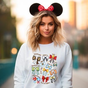 May include: White sweatshirt featuring colorful bow designs inspired by Disney and Pixar movies, including "Ratatouille," "Cars," and "The Incredibles." The model is wearing a Minnie Mouse ear headband with a red polka dot bow.