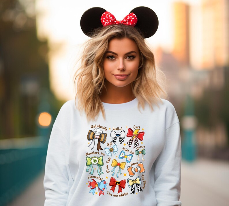 May include: White sweatshirt featuring colorful bow designs inspired by Disney and Pixar movies, including "Ratatouille," "Cars," and "The Incredibles." The model is wearing a Minnie Mouse ear headband with a red polka dot bow.