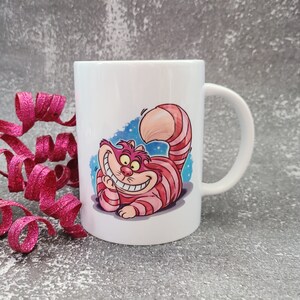 May include: White ceramic mug with a colorful illustration of the Cheshire Cat from Alice in Wonderland. The cat is grinning with a wide, toothy smile and has pink and red stripes.