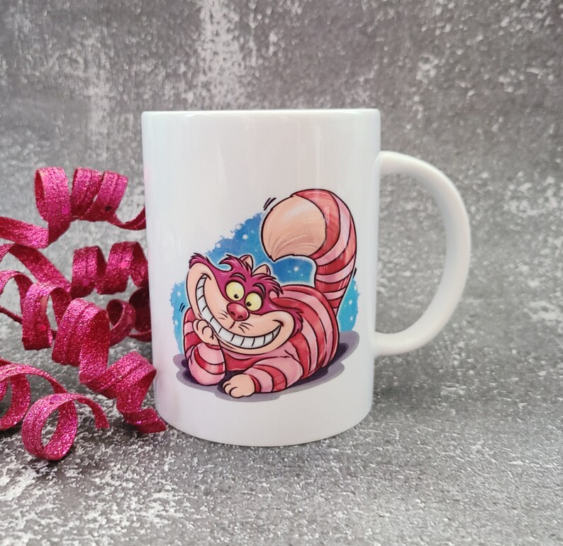 May include: White ceramic mug with a colorful illustration of the Cheshire Cat from Alice in Wonderland. The cat is grinning with a wide, toothy smile and has pink and red stripes.