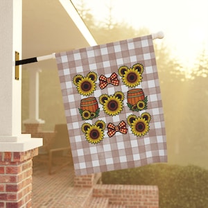 Puede incluir: Una bandera de jardín rectangular con un patrón de cuadros vichy en marrón claro y blanco. El diseño incluye un motivo repetitivo de girasoles con pétalos en forma de orejas de ratón, barriles de madera y lazos a cuadros rojos y blancos.