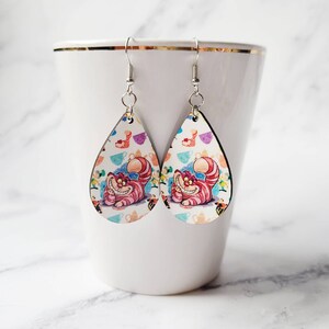 May include: A pair of teardrop-shaped earrings featuring the Cheshire Cat from Alice in Wonderland. The earrings have a white background with colorful teacups and the cat's smiling face. The earrings hang from silver-colored hooks.