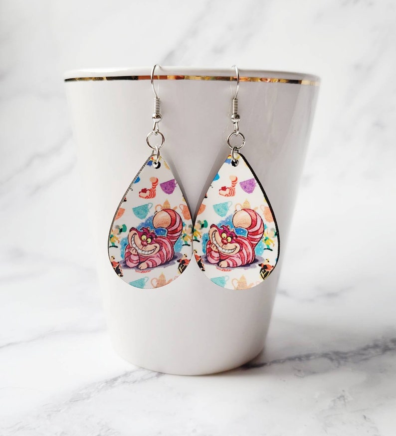 May include: A pair of teardrop-shaped earrings featuring the Cheshire Cat from Alice in Wonderland. The earrings have a white background with colorful teacups and the cat's smiling face. The earrings hang from silver-colored hooks.