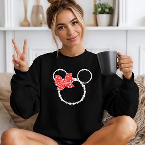 May include: Black sweatshirt featuring a Minnie Mouse design. The design includes a red bow with white polka dots and white pearl-like circles for the ears and head outline. The sweatshirt is a solid black color.