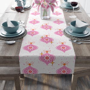 May include: A white table runner featuring a repeating pattern of pink Christmas bauble shapes. The baubles have orange bows and starburst accents. The runner is on a wooden table with place settings and flowers.