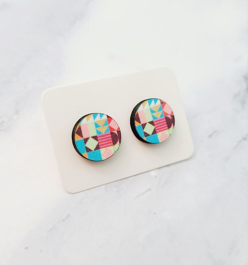 May include: A pair of colourful geometric patterned wooden stud earrings. The earrings are round and have a black backing.