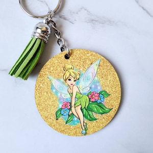 May include: A round wooden keychain with a gold glitter background featuring a cartoon image of Tinkerbell, a fairy with green wings and a green dress, sitting on a leaf with flowers. The keychain has a green tassel attached to a silver ring.