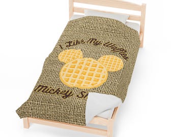 Puede incluir: Una manta de tiro marrón y beige con textura de arpillera. La manta presenta una cabeza de Mickey Mouse hecha de gofres con el texto "Me gustan mis gofres al estilo Mickey".