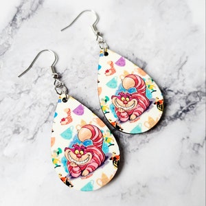 May include: A pair of teardrop-shaped earrings featuring the Cheshire Cat from Alice in Wonderland. The earrings have a white background with colorful teacups and the Cheshire Cat's smiling face. The earrings are made of wood and have silver-colored hooks.