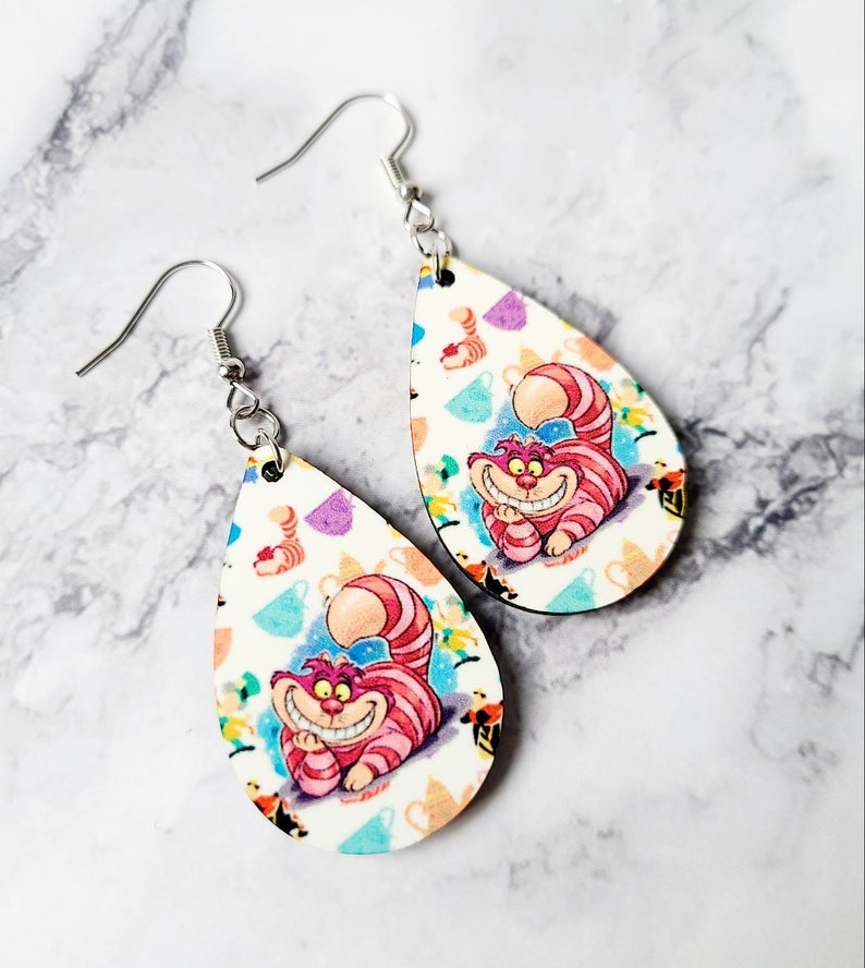 May include: A pair of teardrop-shaped earrings featuring the Cheshire Cat from Alice in Wonderland. The earrings have a white background with colorful teacups and the Cheshire Cat's smiling face. The earrings are made of wood and have silver-colored hooks.