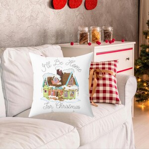 Może przedstawiać: Biała kwadratowa poduszka z kolorową ilustracją domu pokrytego śniegiem, z napisem "I'll Be Home for Christmas". Za nią czerwono-biała poduszka w kratę.