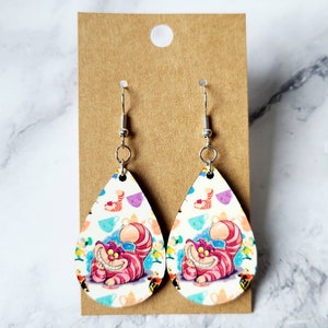 May include: Pair of teardrop-shaped earrings featuring the Cheshire Cat from Alice in Wonderland. The earrings have a white background with colorful teacups and the Cheshire Cat's smiling face. The earrings hang from silver-colored hooks.