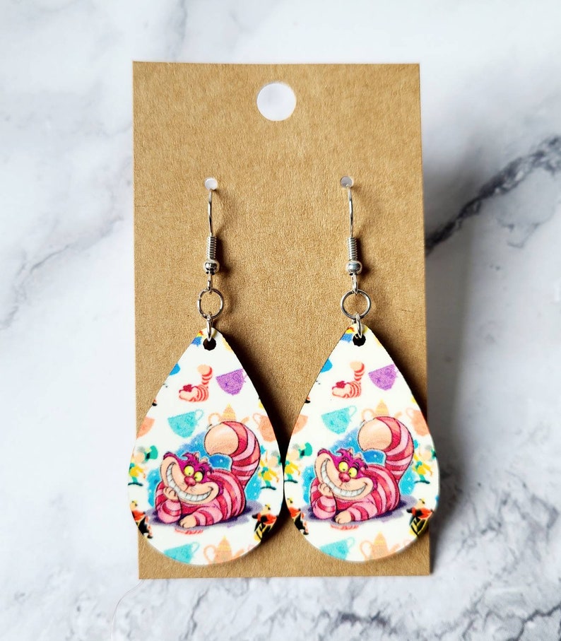 May include: Pair of teardrop-shaped earrings featuring the Cheshire Cat from Alice in Wonderland. The earrings have a white background with colorful teacups and the Cheshire Cat's smiling face. The earrings hang from silver-colored hooks.