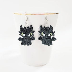 May include: A pair of black glitter dangle earrings featuring a cartoon dragon design with green eyes.