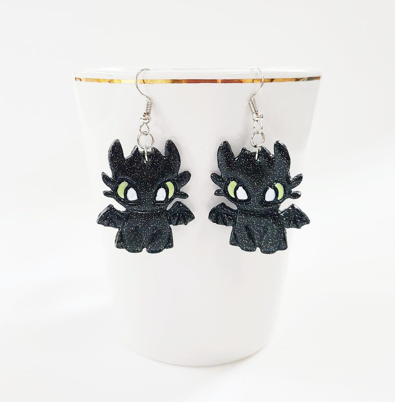 May include: A pair of black glitter dangle earrings featuring a cartoon dragon design with green eyes.