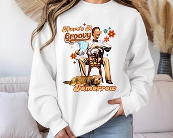 May include: White sweatshirt with a retro graphic. The design features a seated figure with sunglasses, a dog, and the text "There's A Groovy Tomorrow" in brown and orange. The graphic includes flowers and stars.