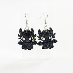 May include: A pair of black glitter dangle earrings featuring a cartoon dragon design with green eyes.