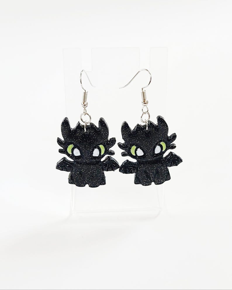 May include: A pair of black glitter dangle earrings featuring a cartoon dragon design with green eyes.