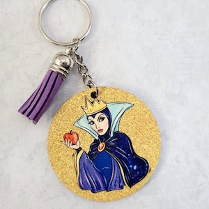 May include: A round wooden keychain with a gold glitter background featuring a cartoon image of the Evil Queen from Snow White holding a red apple. The Evil Queen is wearing a purple and blue gown, a crown, and a cape. The keychain has a purple tassel attached to a silver ring.