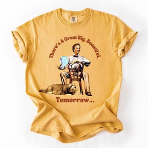May include: Mustard yellow t-shirt featuring a vintage-style illustration. The graphic depicts a man seated in a chair with a dog at his feet. Text on the shirt reads "There's A Great Big, Beautiful, Tomorrow..."