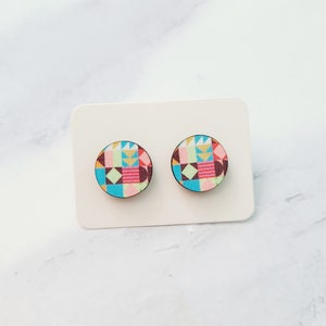 May include: A pair of colourful geometric patterned wooden earrings. The earrings are round and have a light blue background with pink, yellow, green, and brown geometric shapes.