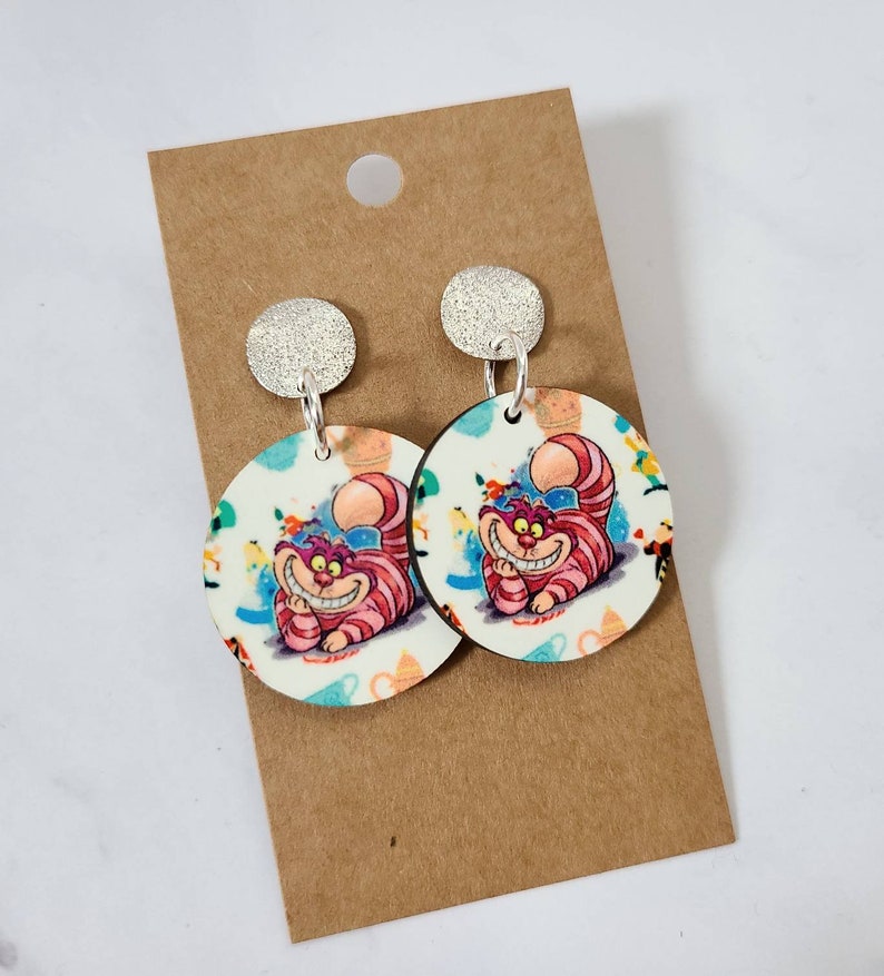May include: Round dangle earrings featuring the Cheshire Cat from Alice in Wonderland. The earrings have a silver glitter top and a circular design with the cat's smiling face in pink and white stripes. The background has colorful illustrations.