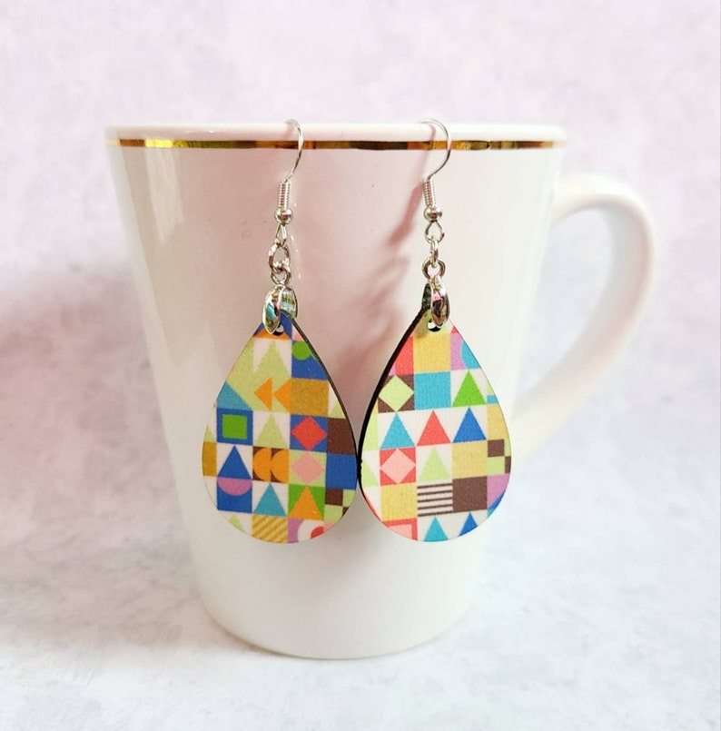 May include: A pair of teardrop-shaped earrings with a colourful geometric pattern. The earrings are made of wood and have silver hooks.