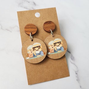 May include: A pair of wooden earrings featuring a cartoon image of an elderly couple. The earrings are brown and have a round shape. The couple is depicted sitting at a table and holding mugs.