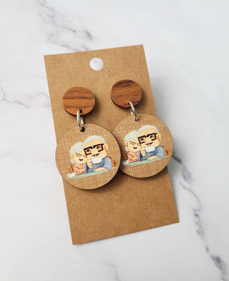 May include: A pair of wooden earrings featuring a cartoon image of an elderly couple. The earrings are brown and have a round shape. The couple is depicted sitting at a table and holding mugs.