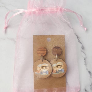 May include: A pair of wooden earrings with a cartoon image of an older couple. The earrings are packaged in a pink organza bag.