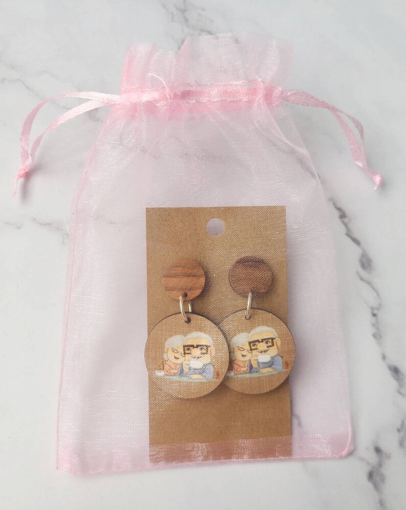 May include: A pair of wooden earrings with a cartoon image of an older couple. The earrings are packaged in a pink organza bag.