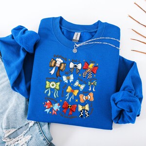 May include: A royal blue sweatshirt featuring a collection of colorful bow tie illustrations. The bow ties are designed with various patterns and colors, and some include text. A silver necklace with a pendant is also visible.