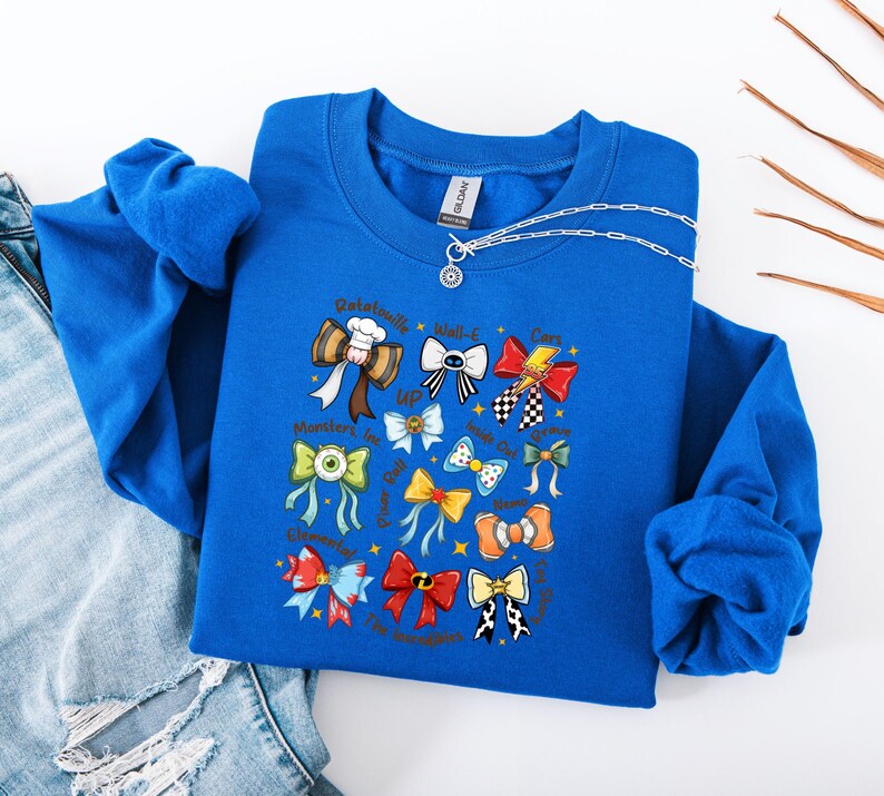May include: A royal blue sweatshirt featuring a collection of colorful bow tie illustrations. The bow ties are designed with various patterns and colors, and some include text. A silver necklace with a pendant is also visible.