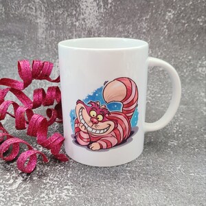 May include: White ceramic mug with a colorful illustration of the Cheshire Cat from Alice in Wonderland. The cat is grinning and has pink and white stripes.