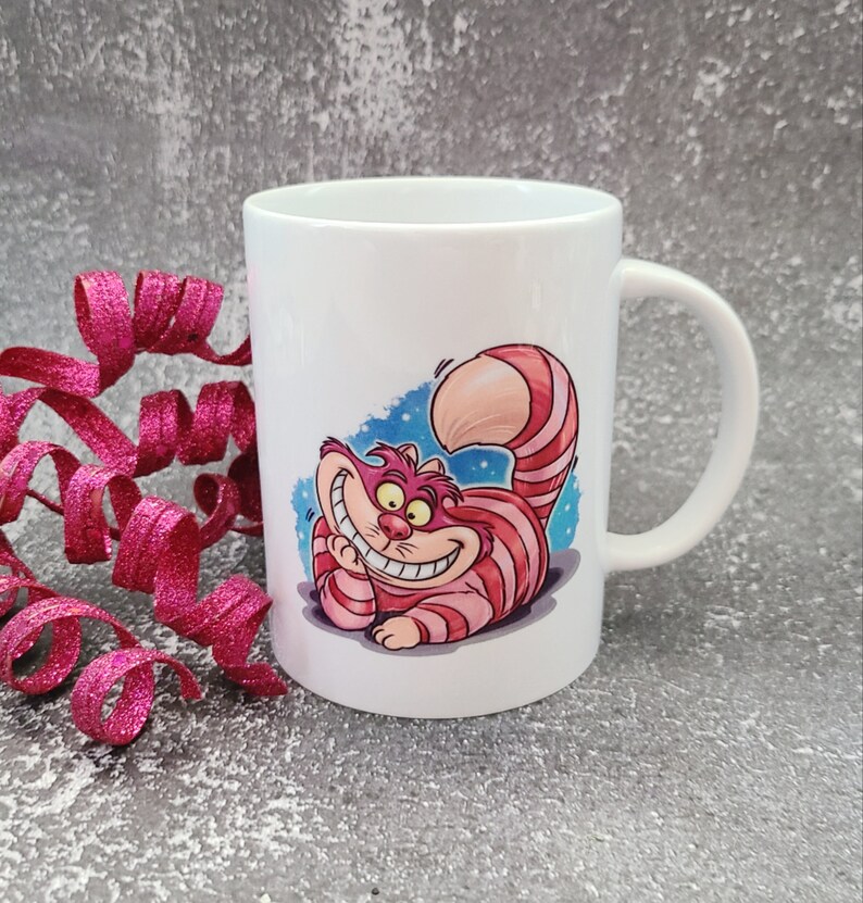 May include: White ceramic mug with a colorful illustration of the Cheshire Cat from Alice in Wonderland. The cat is grinning and has pink and white stripes.