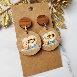 May include: A pair of wooden earrings with a round, brown wood top and a round, printed image of an older couple. The image is of a man and woman sitting at a table, smiling and holding mugs. The earrings are on a brown card.