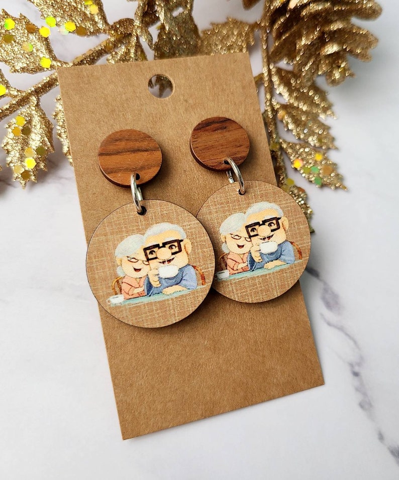 May include: A pair of wooden earrings with a round, brown wood top and a round, printed image of an older couple. The image is of a man and woman sitting at a table, smiling and holding mugs. The earrings are on a brown card.