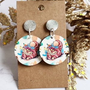 May include: A pair of round earrings featuring the Cheshire Cat from Alice in Wonderland. The earrings have a silver stud and a white background with colorful illustrations of the cat and teacups. The cat is pink and purple with striped details.
