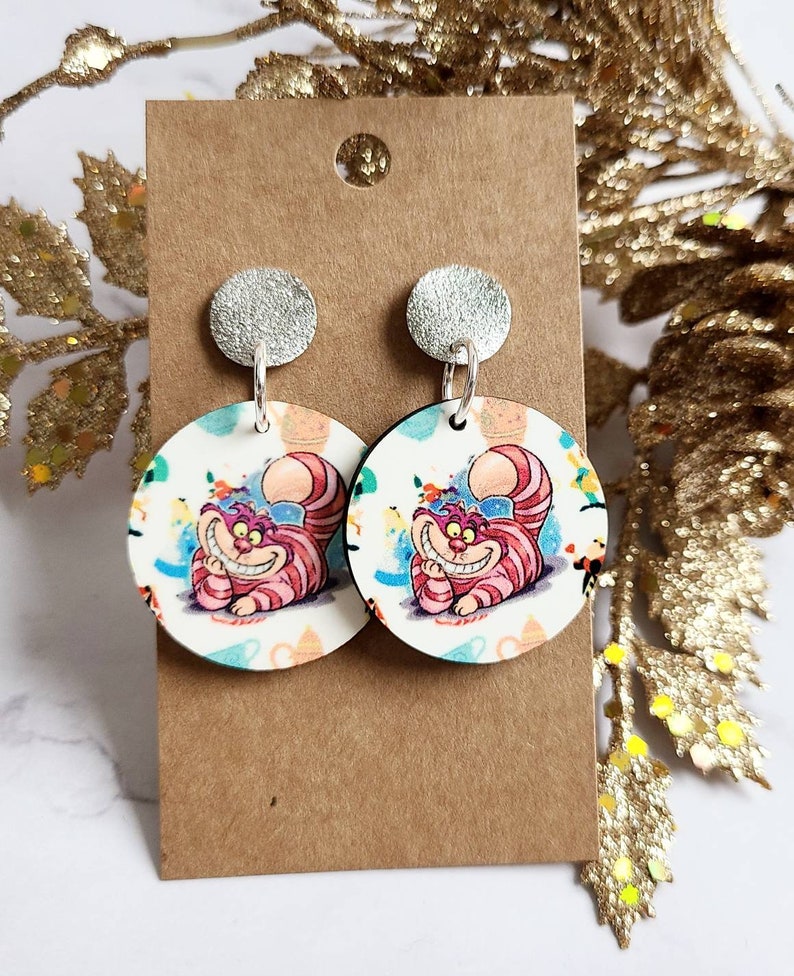 May include: A pair of round earrings featuring the Cheshire Cat from Alice in Wonderland. The earrings have a silver stud and a white background with colorful illustrations of the cat and teacups. The cat is pink and purple with striped details.