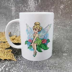 May include: White ceramic mug with a colorful illustration of Tinkerbell, a fairy with green wings, sitting on a leaf with flowers.