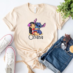 May include: A beige t-shirt with a cartoon image of Stitch and Lilo from the Disney movie Lilo & Stitch. Stitch is hugging Lilo and the text "Ohana" is printed below the image.
