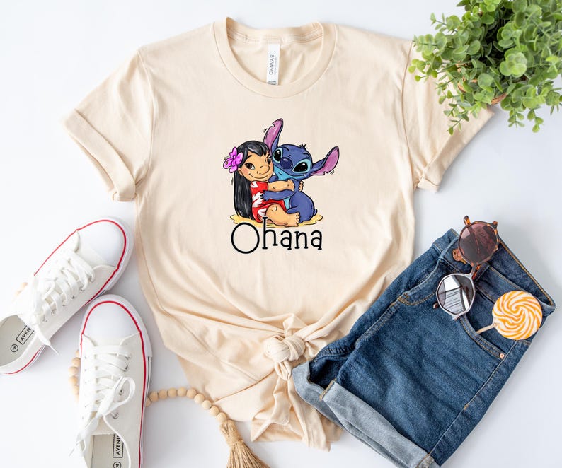 May include: A beige t-shirt with a cartoon image of Stitch and Lilo from the Disney movie Lilo & Stitch. Stitch is hugging Lilo and the text "Ohana" is printed below the image.