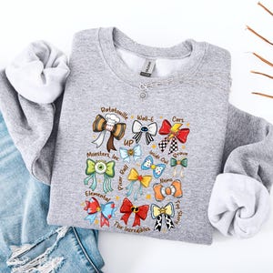 May include: A gray sweatshirt featuring colorful bow designs representing various animated movies, including "Ratatouille," "Wall-E," and "Cars." The sweatshirt has a crew neck and long sleeves. The text "GILDAN" is visible on the tag.