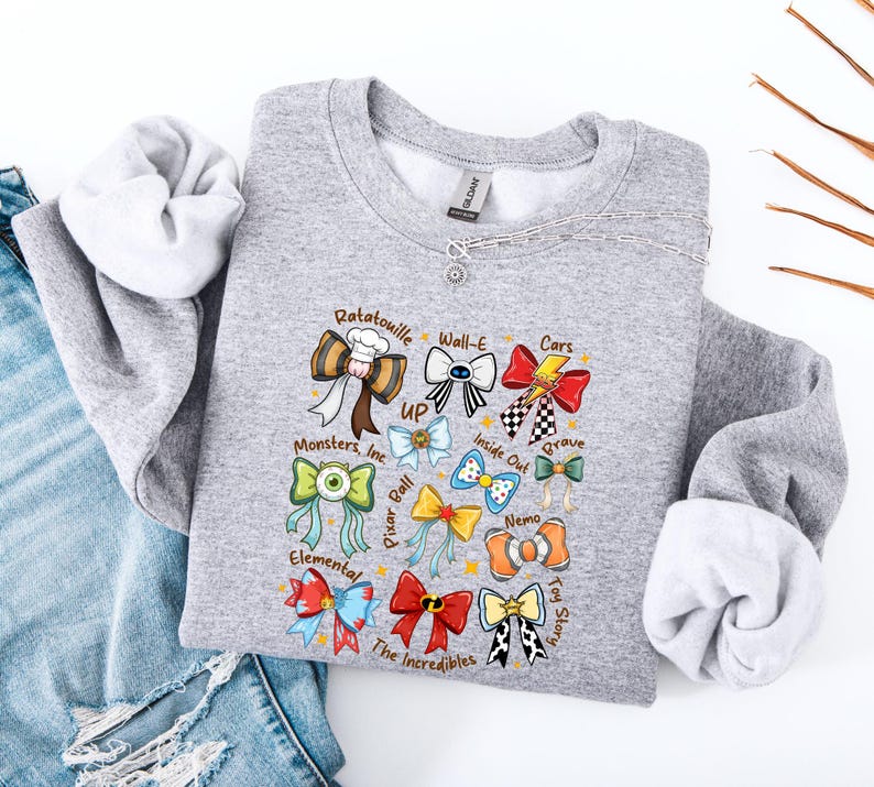 May include: A gray sweatshirt featuring colorful bow designs representing various animated movies, including "Ratatouille," "Wall-E," and "Cars." The sweatshirt has a crew neck and long sleeves. The text "GILDAN" is visible on the tag.